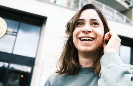 A smiling woman with long brown hair is wearing a green sweatshirt, holding a phone to her ear outside a building. The background shows a slightly blurred facade with glass doors and railings.