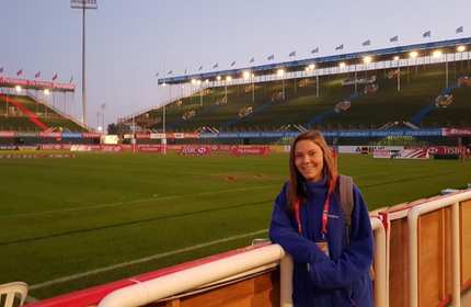 Sophie Scherschel leans on the back of the advertising boards looking over a rugby stadium pitch and stands