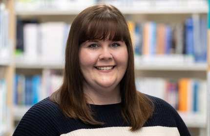 A close-up of a person smiling. In the background are some library bookshelves.
