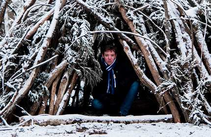 A person wearing a scarf sits inside a makeshift shelter made of branches in a snowy forest.