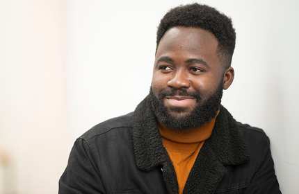 Ted Airaga, wearing an orange turtleneck and black jacket, leans against a white wall