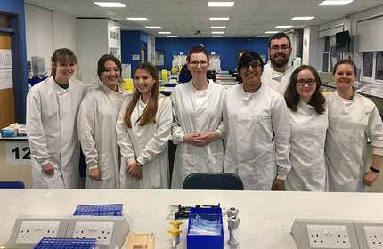 A group of eight people wearing white lab coats stands in a laboratory. They are smiling and posing for the photo, with lab equipment and workstations visible in the background.