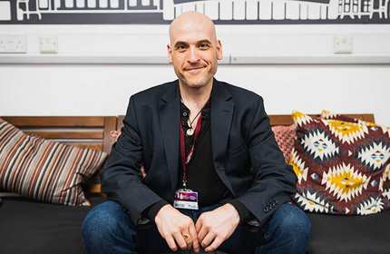 A bald man wearing a black blazer is sitting on a dark sofa. He is smiling and has his hands clasped. The sofa has various patterned cushions. The background features a black and white wall design.