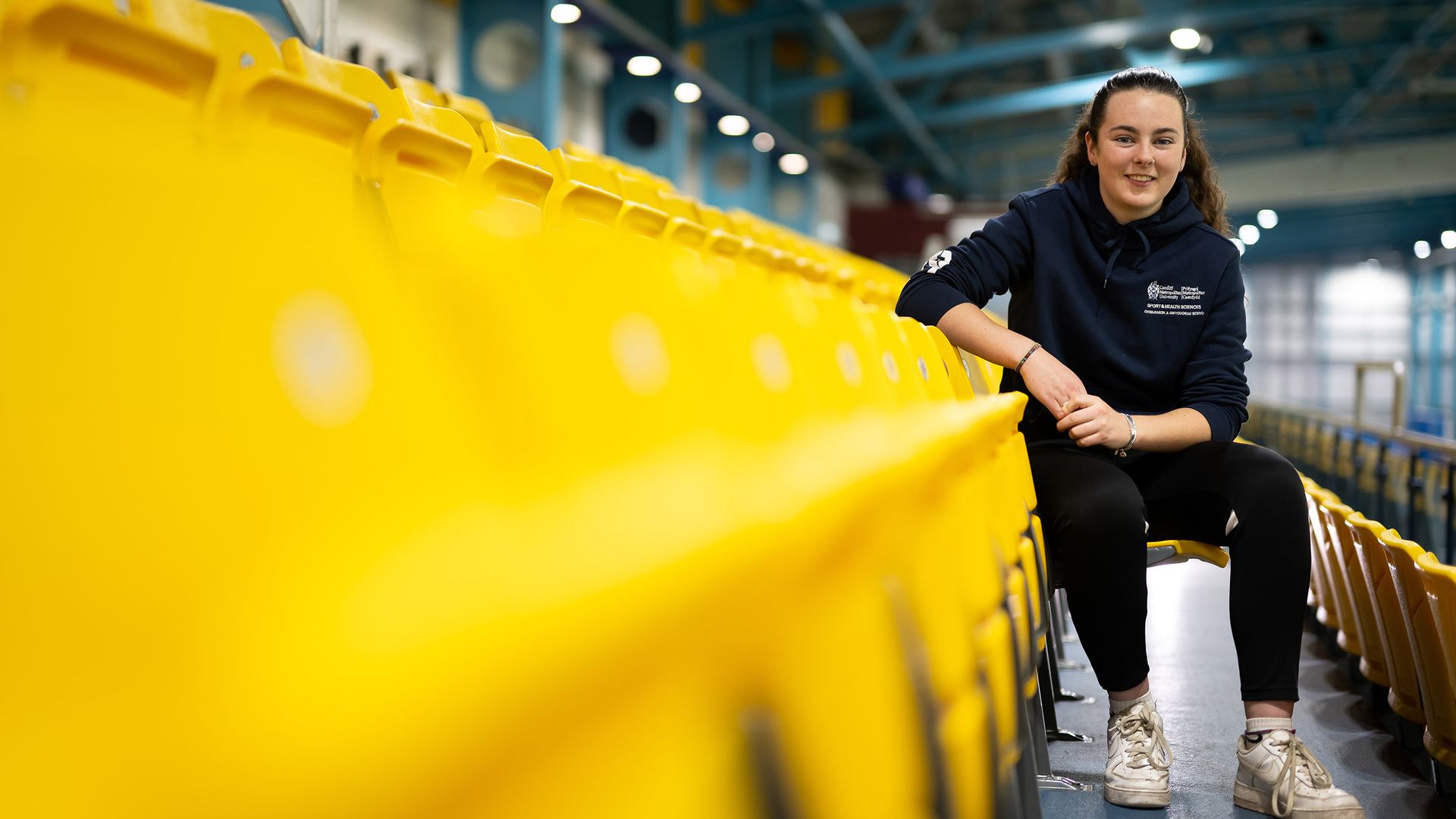 A woman with long dark hair sits on yellow bleachers in an indoor sports facility. She is wearing a black T-shirt, blue jeans, and white sneakers, and is smiling while looking to the side. The background features a running track.