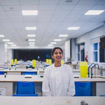A woman with long hair stands confidently in a medical examination room, arms crossed. Behind her is a model skeleton, medical equipment, and a bed. She is wearing a dark cardigan over a white shirt and smiling.