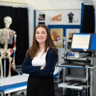 A person in a white lab coat stands smiling in a bright, modern laboratory with blue chairs and yellow safety containers on the tables. The room is well-lit with numerous ceiling lights.