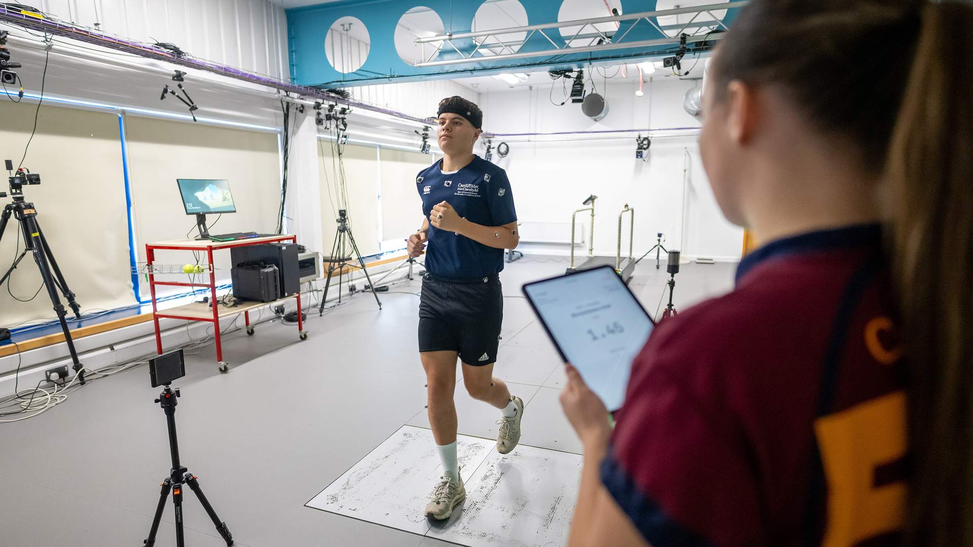 A person is jogging indoors in a lab setup with various sensors and equipment. Another individual holds a tablet, observing the activity. The room has a high ceiling and multiple cameras and lights are mounted around.