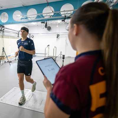 A person is jogging indoors in a lab setup with various sensors and equipment. Another individual holds a tablet, observing the activity. The room has a high ceiling and multiple cameras and lights are mounted around.