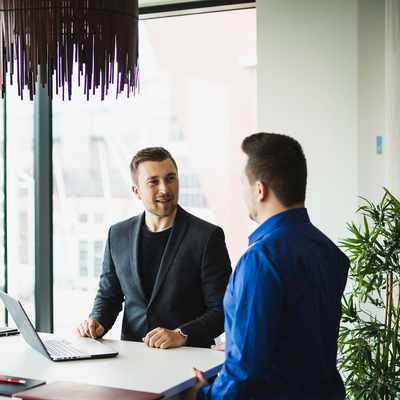 Two men are seated at a white table in a modern office with large windows. One is using a laptop, and they appear to be having a conversation. Natural light fills the room, and theres a potted plant nearby.