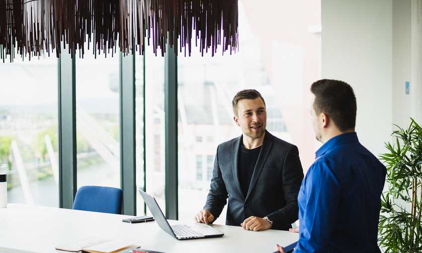 Two men are seated at a white table in a modern office with large windows. One is using a laptop, and they appear to be having a conversation. Natural light fills the room, and theres a potted plant nearby.