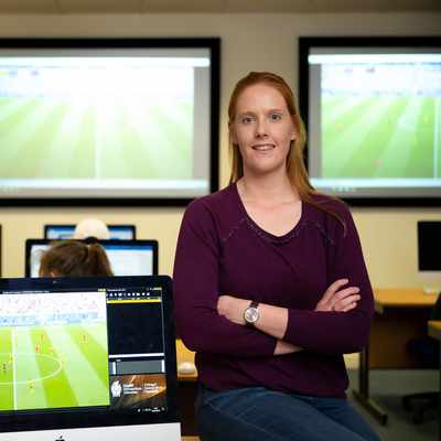 A woman with long red hair, wearing a purple sweater, stands confidently with arms folded in a classroom. People are working at computers, and a soccer game is projected on two large screens in the background.