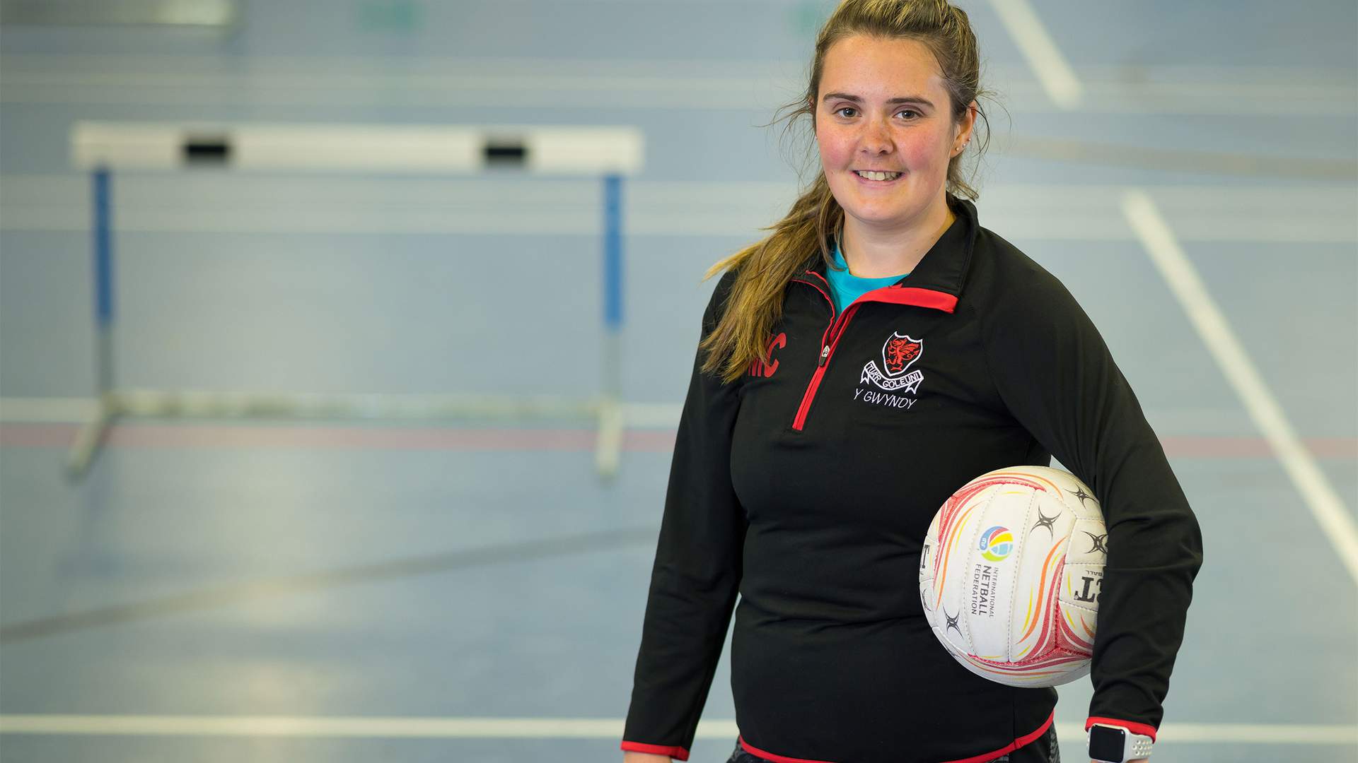 A person in sports attire stands in an indoor court, holding a netball with one hand. The court has visible markings and a hurdle in the background. The person is smiling and wearing a black jacket with a logo.