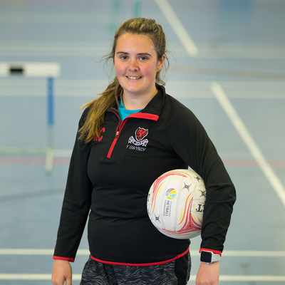 A person in sports attire stands in an indoor court, holding a netball with one hand. The court has visible markings and a hurdle in the background. The person is smiling and wearing a black jacket with a logo.