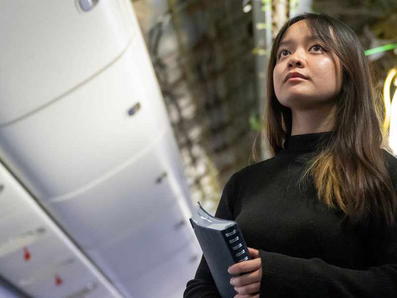 A woman stands inside an airplane, holding a ring binder, inspecting the wiring above.
