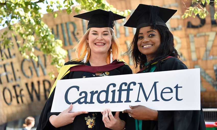 Two Cardiff Met students in graduation caps and gowns holding a Cardiff Met sign
