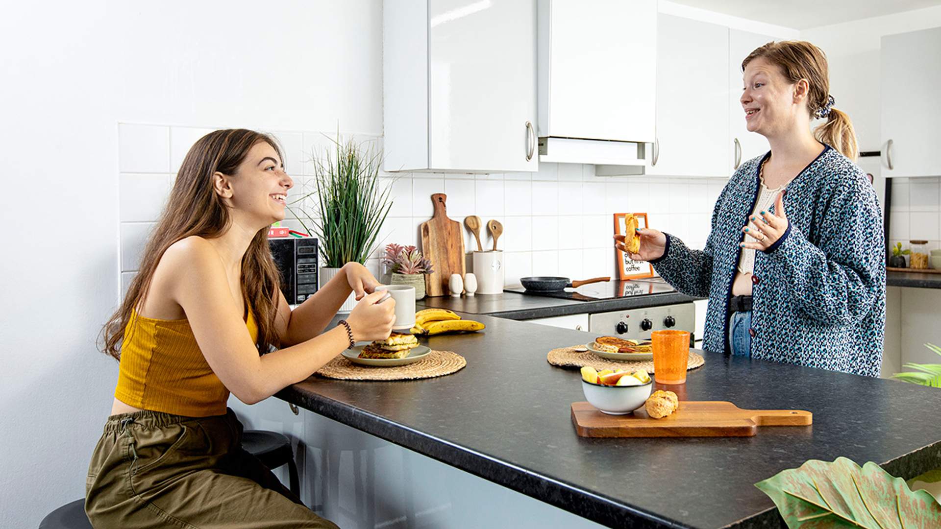 Two female students engaged in conversation while sitting at a kitchen counter, surrounded by kitchen items and warm lighting.