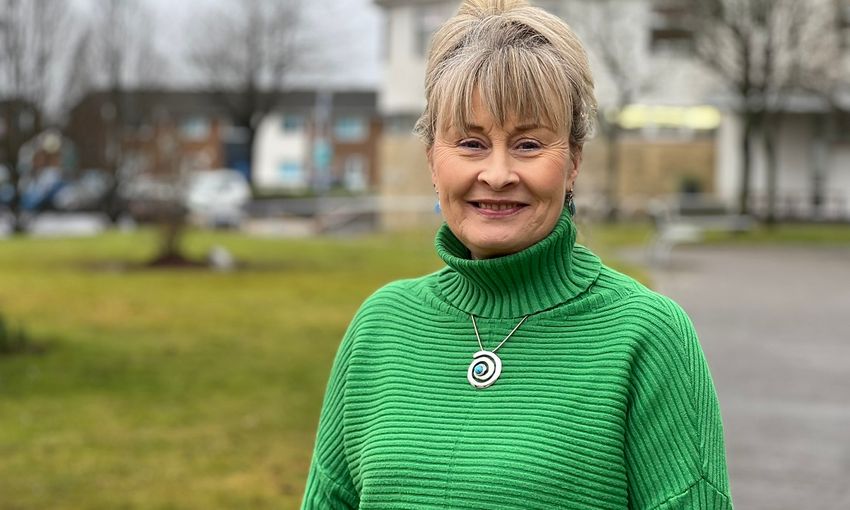A woman wearing a green sweater stands confidently in front of a building, showcasing her stylish attire and urban backdrop.