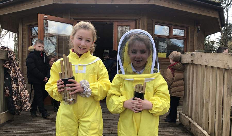 Two children wearing beekeeping suits stand in front of a small wooden building.