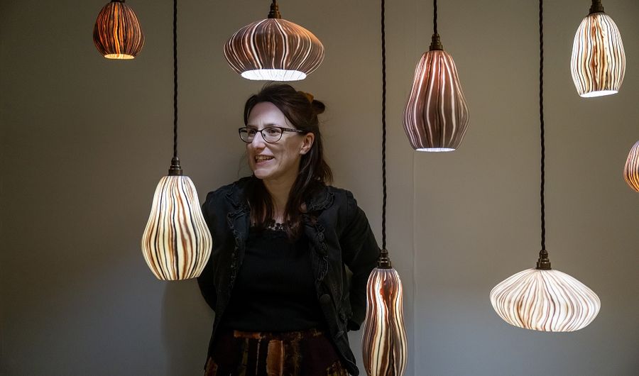 A woman wearing glasses leans against a white wall behind a collection of hanging lights