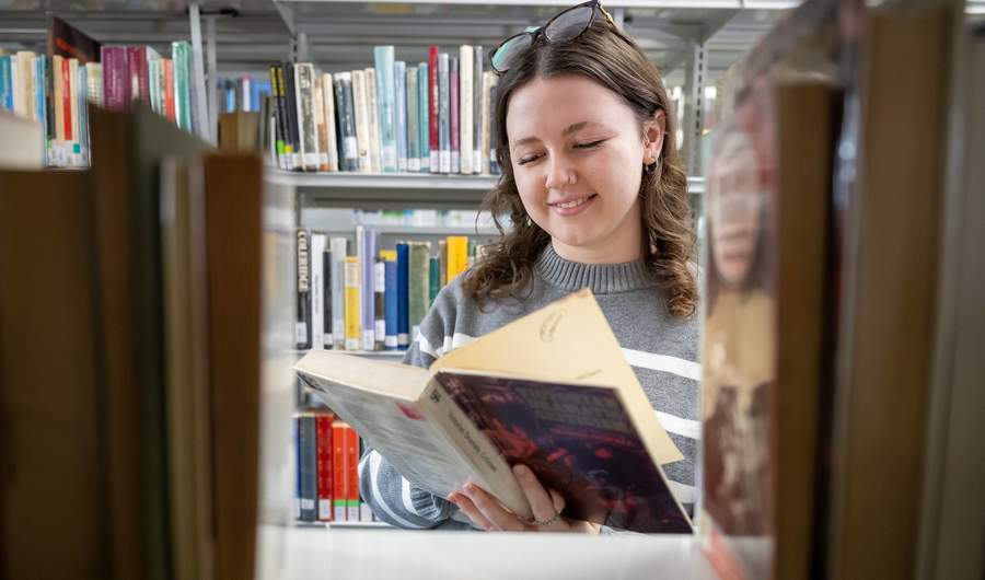 A young adult reads a book in between the bookshelves at one of the on-campus libraries