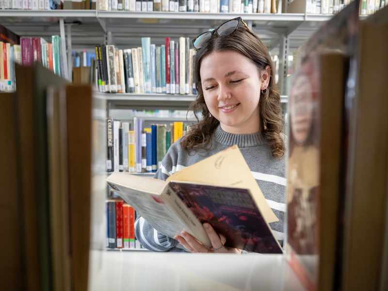 A young adult reads a book in between the bookshelves at one of the on-campus libraries