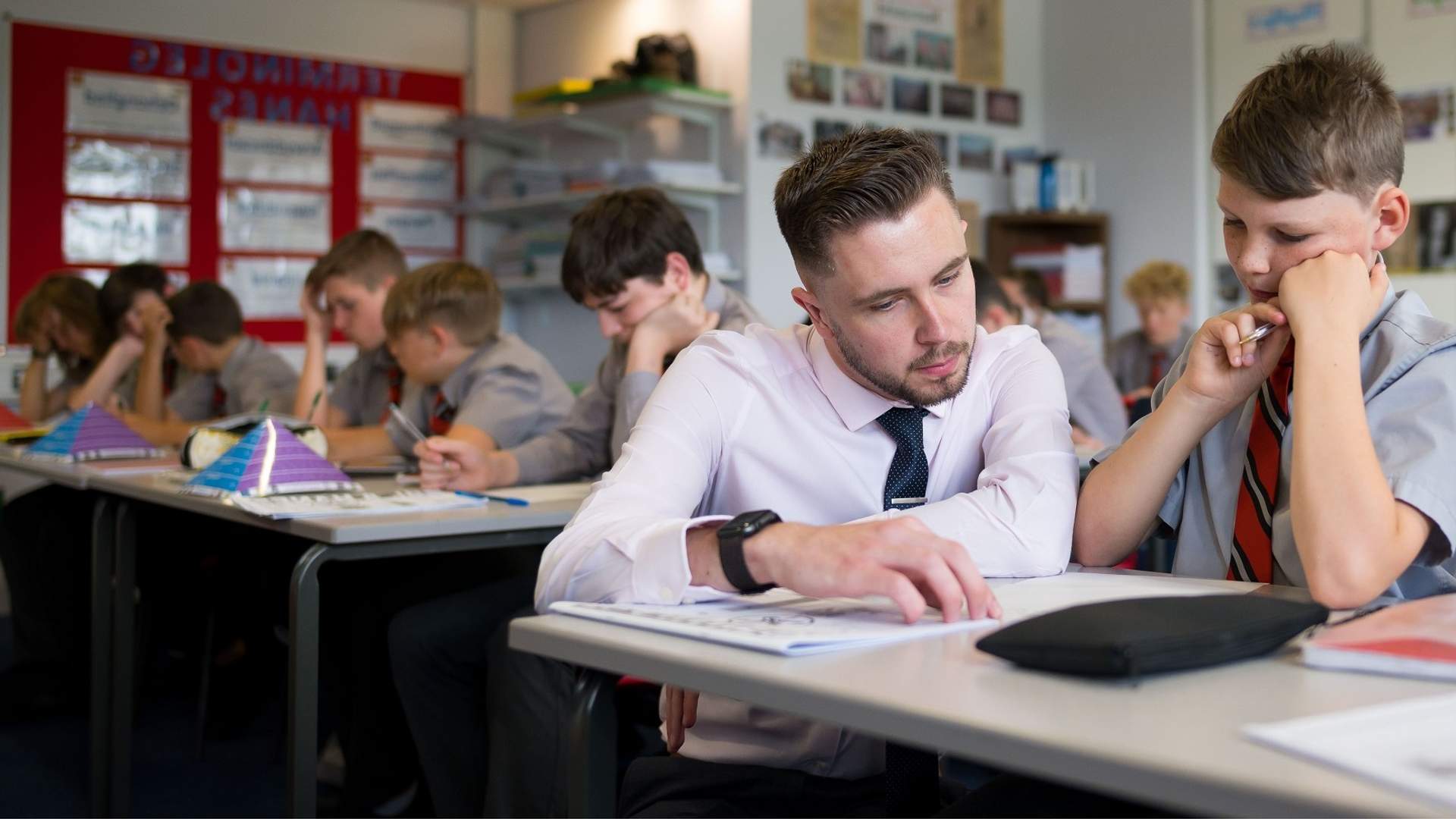 A teacher kneels down to help a pupil working through a booklet at their classroom desk