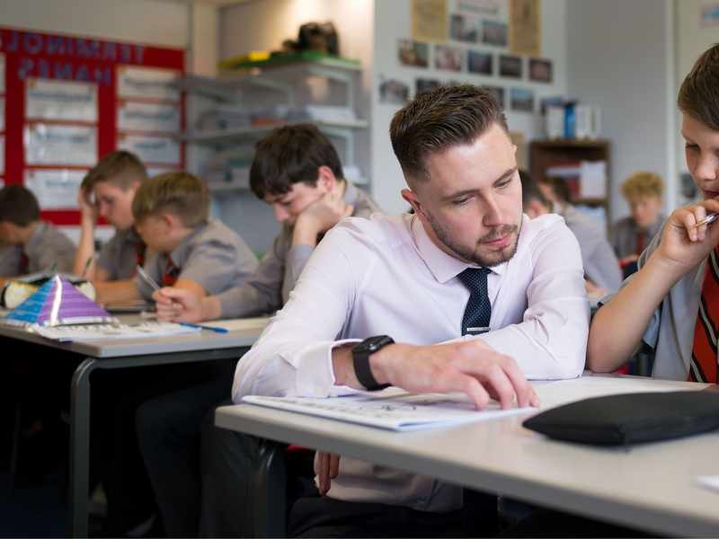 A teacher kneels down to help a pupil working through a booklet at their classroom desk
