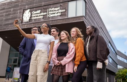 A group of six young adults take a selfie in front of a building with the sign Cardiff Metropolitan University. They are smiling and appear to be enjoying their time together. The sky is partly cloudy.
