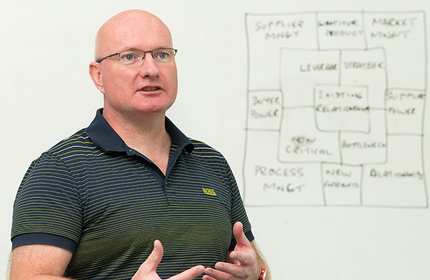 Man wearing a polo t-shirt and glasses engages in discussion in front of a whiteboard