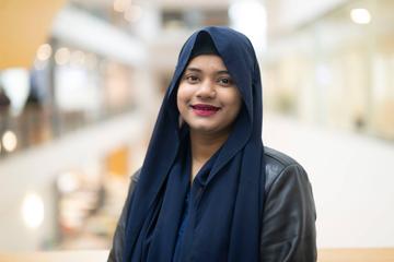 A person wearing a blue headscarf and black leather jacket stands in a bright atrium space.