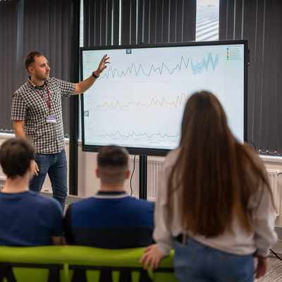 A person stands in front of a small group of people, pointing to charts on a large screen.