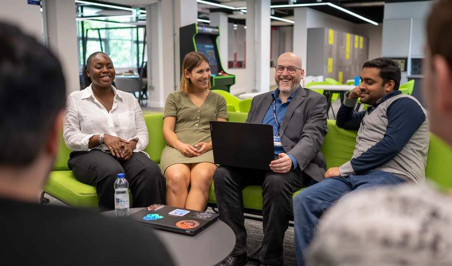 A group of people sit around a small round table. One person has an open laptop computer on their lap.