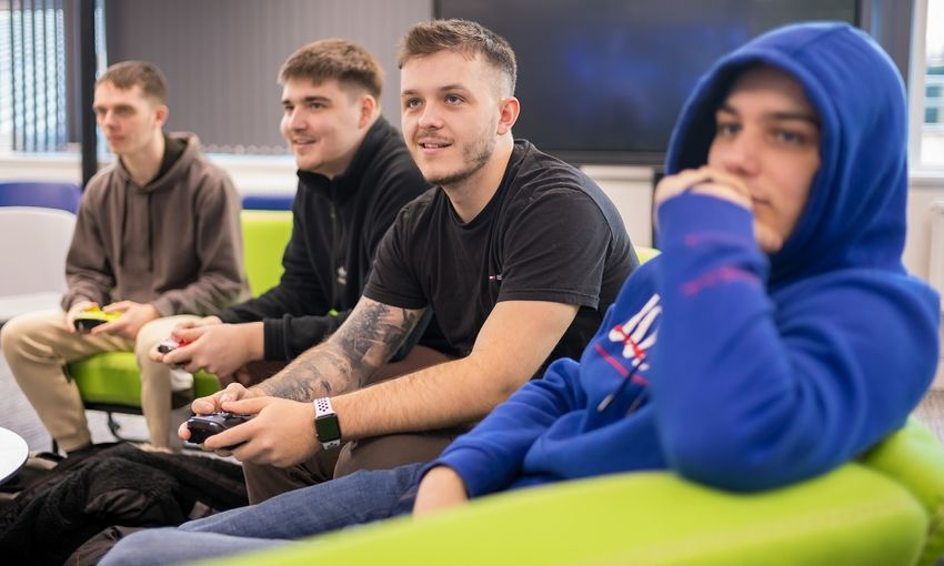 Four students sit on a curved green sofa. One is holding a gaming controller and they are all looking in the same direction