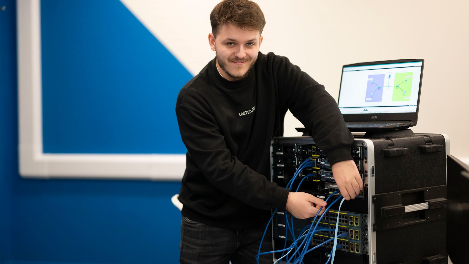 A young adult smiles for the camera as they work on a computer tower and adjust the attached cables