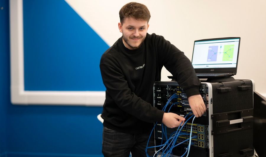 A young adult smiles for the camera as they work on a computer tower and adjust the attached cables