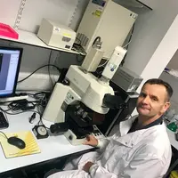 A man in a lab coat sits at a desk, focused on a microscope, surrounded by scientific equipment and notes.