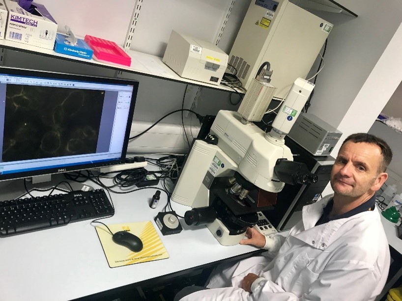 A man in a lab coat sits at a desk, focused on a microscope, surrounded by scientific equipment and notes.