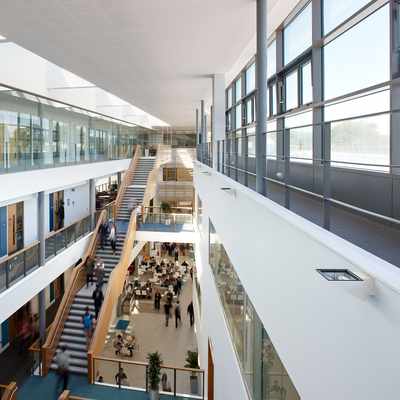 A modern, spacious atrium with a large wooden staircase and multiple people walking. The area has glass railings, plants, and abundant natural light from skylights above. The scene conveys a busy, contemporary office or educational environment.