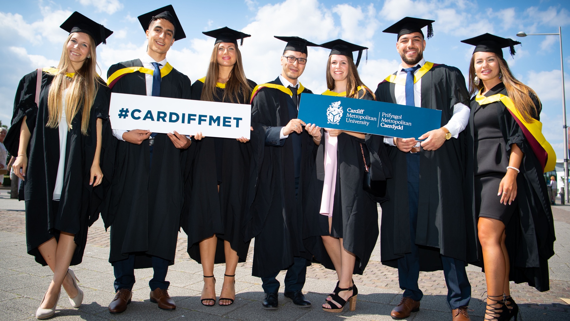 A group of students in graduation caps and gowns stand holding Cardiff Met signs