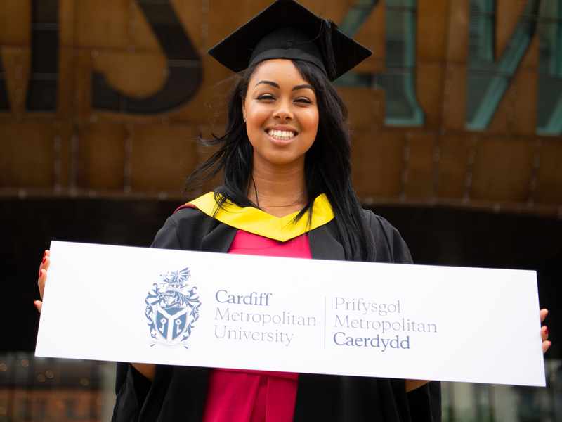 A Cardiff Met student, in graduation cap and gown, stands holding a sign featuring the Cardiff Met logo and name