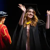 A smiling graduate in gown and cap raises her arms in celebration on stage during the Cardiff Metropolitan University Class of 2025 graduation ceremony, as a university official in red and gold academic robes looks on.