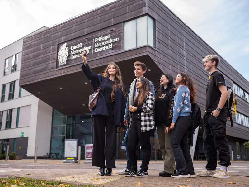 Six young adults stand huddled together outside the Cardiff School of Management as one of them takes a selfie on a mobile phone.