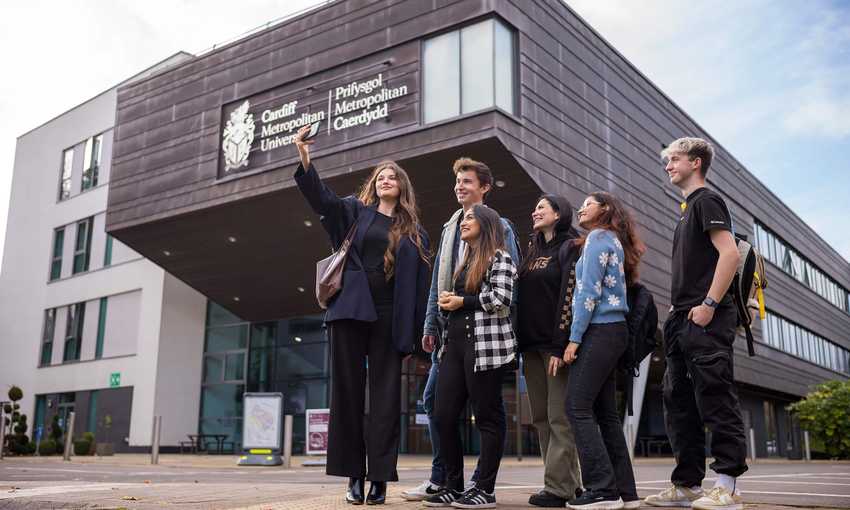 Six young adults stand huddled together outside the Cardiff School of Management as one of them takes a selfie on a mobile phone.