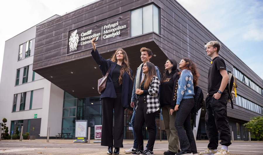 Six young adults stand huddled together outside the Cardiff School of Management as one of them takes a selfie on a mobile phone.