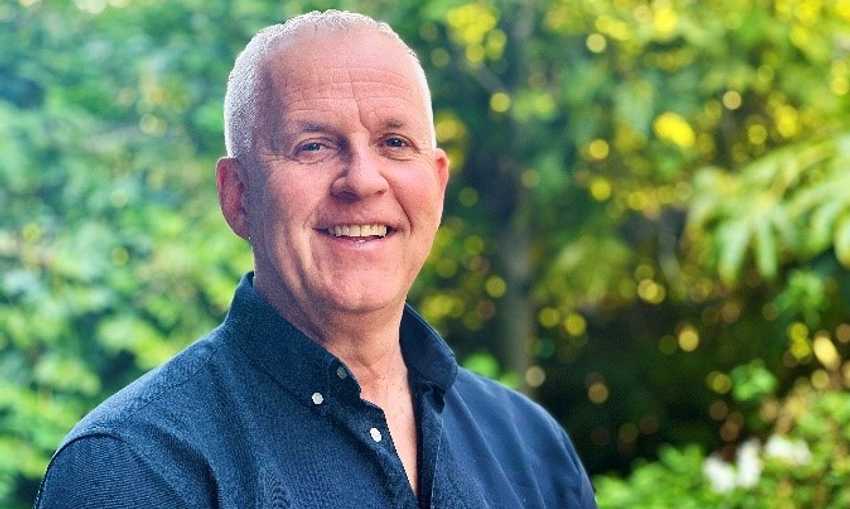 A man in a blue shirt smiles warmly in front of a backdrop of lush green trees.