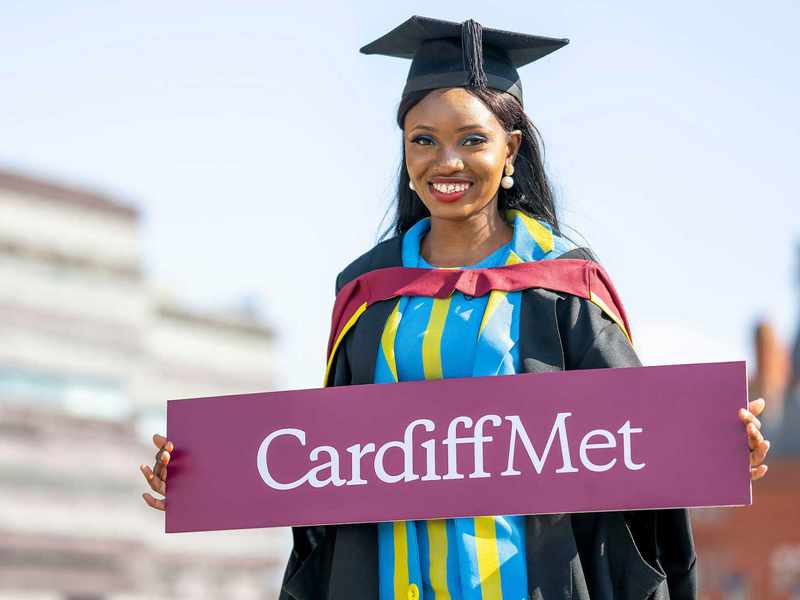A woman in a graduation gown proudly holds a sign that reads 