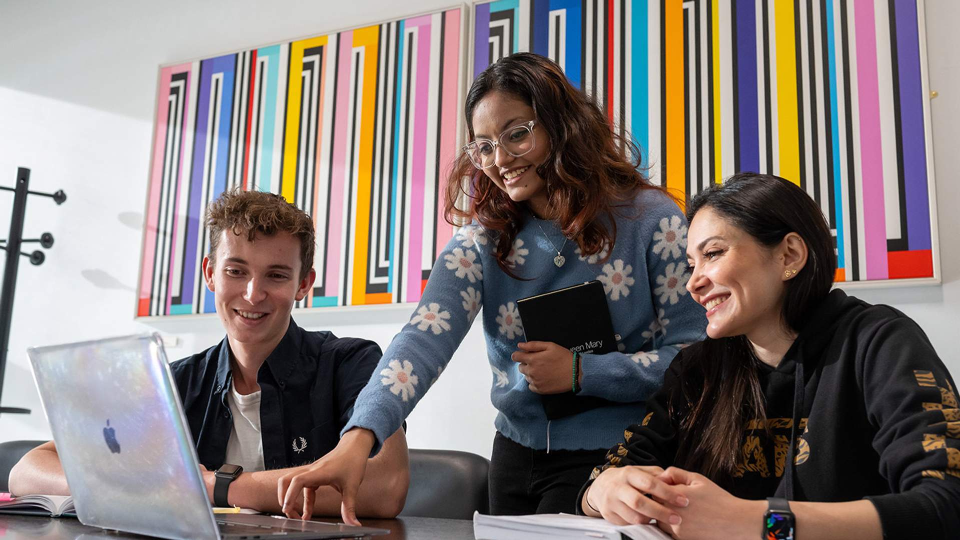 Three students collaborating on a laptop in a modern office setting, focused on their project.