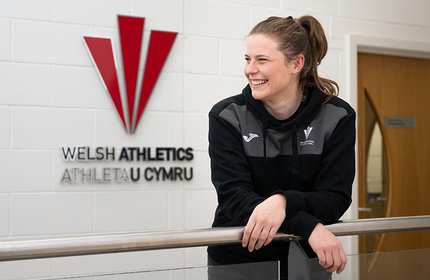 Jana Pacyna leans forward on a railing, in front of the Welsh Athletics logo