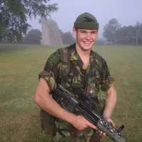 A man in camouflage clothing holds a rifle, positioned in a wooded area, indicating readiness and focus.