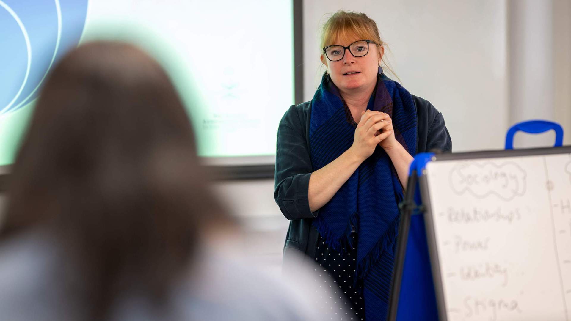 A woman wearing glasses stands in front of a whiteboard, engaged in a discussion or presentation.